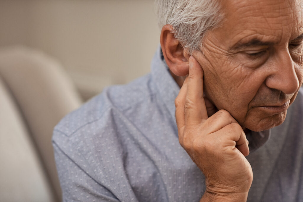 Senior man with hearing problems Side view of senior man with symptom of hearing loss. Mature man sitting on couch with fingers near ear suffering pain.