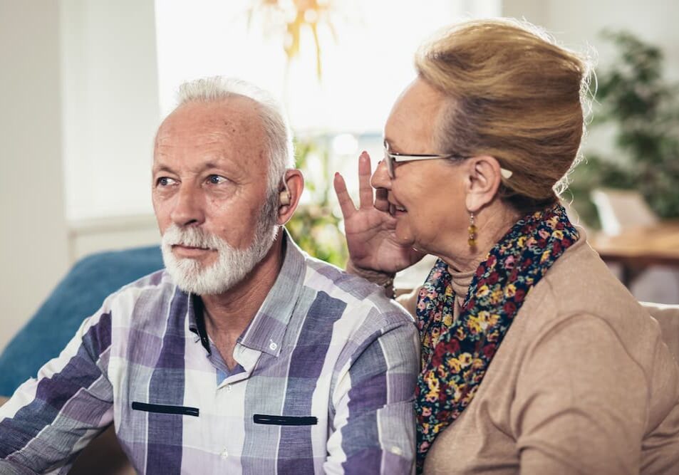 Wife speaking into husband's ear, as he adjusts to new hearing aids.